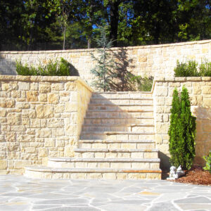 Stone staircase leading up to a raised garden area with light-colored stone walls and greenery. A small conifer tree is placed at the top of the stairs. The foreground features a paved stone courtyard. StoneSupplyStore.com We Sell Stone, Repair Stone, and Do Stone Projects